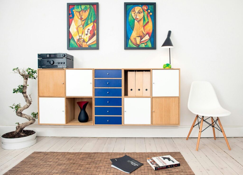 Modern living room corner featuring a wooden cabinet with compartments and drawers, topped with a stereo system, a black lamp, a small green plant, and two colorful abstract paintings above. A bonsai tree in a white pot sits to the left, and a white chair with wooden legs is on the right. Two open books lie on a woven rug in front of the cabinet.
