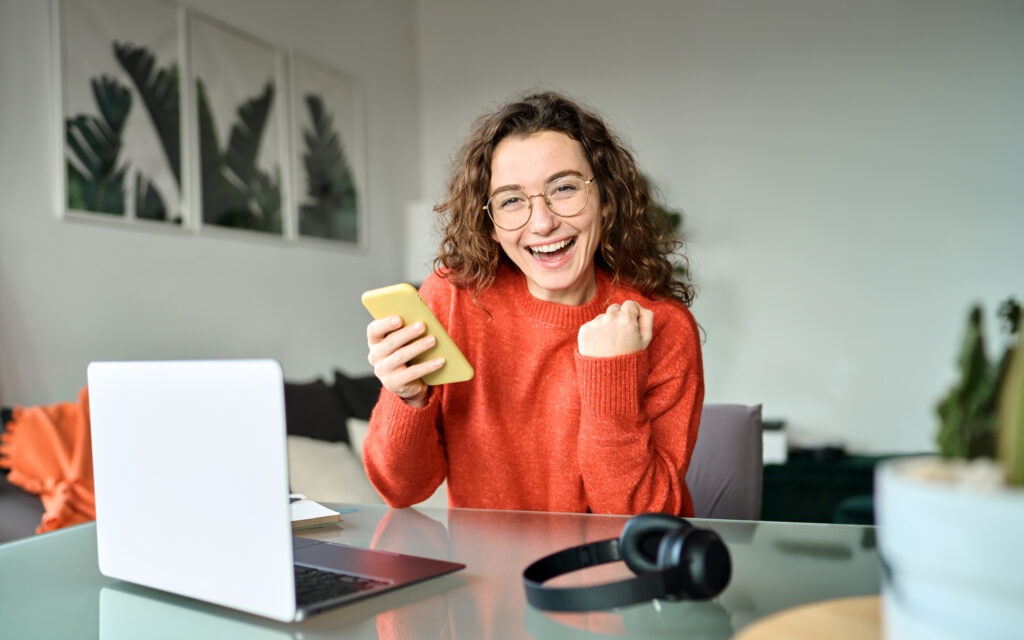 A person with curly hair, wearing an orange sweater, is sitting at a desk with a laptop and holding a yellow smartphone. The person's face is blurred. On the desk, there are also headphones and some papers. In the background, there are three framed pictures of leaves on the wall.
