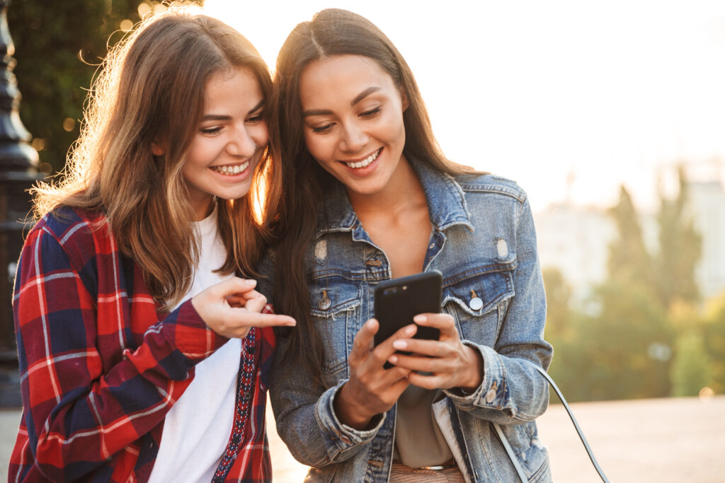 Two individuals are standing outdoors, looking at a smartphone. The individual on the left is wearing a red and blue plaid shirt over a white top and is pointing at the phone. The individual on the right is wearing a denim jacket and holding the phone with both hands. Their faces are obscured by pixelation. The background shows trees and sunlight.