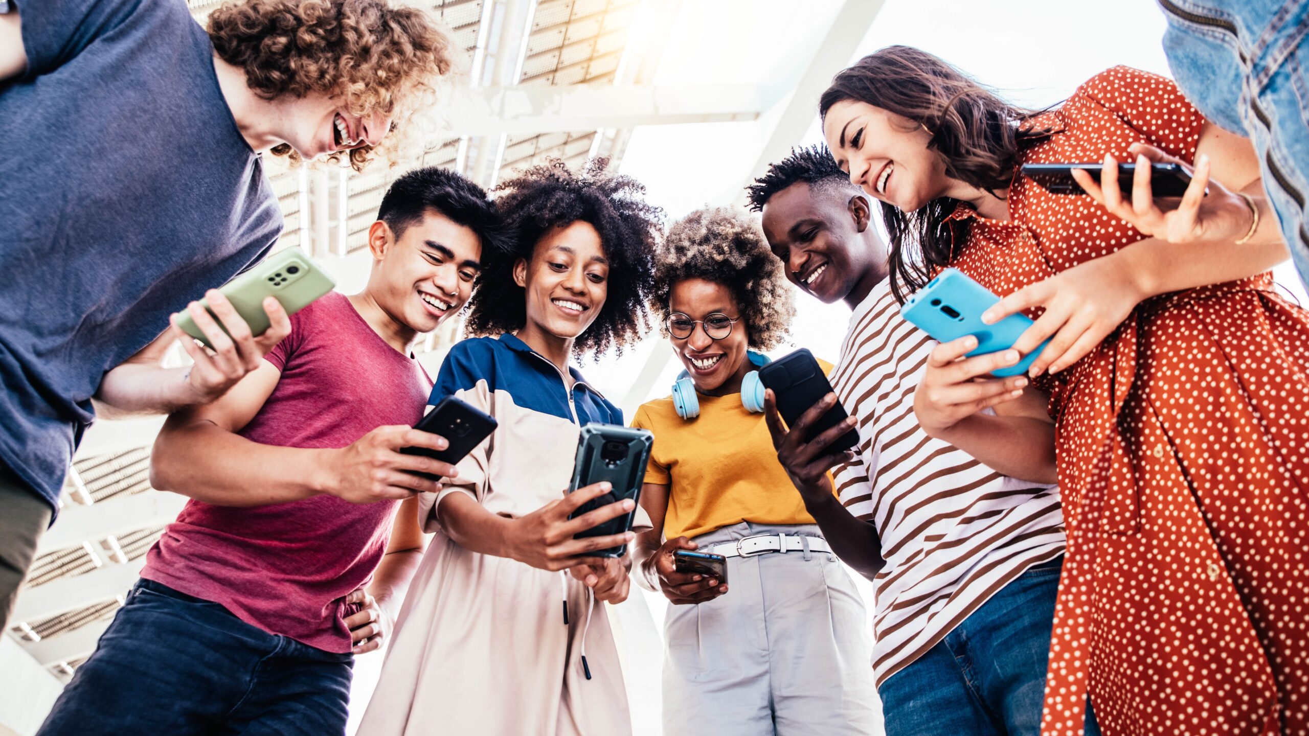 A group of people standing in a circle, each holding and looking at their smartphones. The individuals are dressed in casual clothing and appear to be outdoors under a structure with a glass roof. The focus is on the smartphones they are holding.
