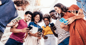 A group of people standing in a circle, each holding and looking at their smartphones. The individuals are dressed in casual clothing and appear to be outdoors under a structure with a glass roof. The focus is on the smartphones they are holding.