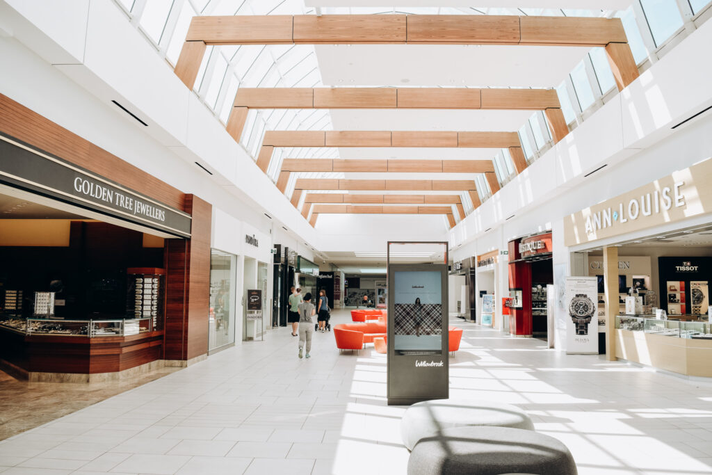 Interior view of Willowbrook Shopping Centre in Langley, BC, featuring stores like Golden Tree Jewellers and Ann-Louise, with high wooden-beam ceilings, skylights, red seating areas, and an information kiosk.
