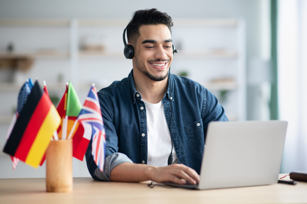 A person wearing a headset is sitting at a desk with a laptop. On the desk, there is a small wooden container holding miniature flags of various countries, including Germany, Italy, and the United Kingdom. The background shows shelves with some items on them and a window with natural light coming in.