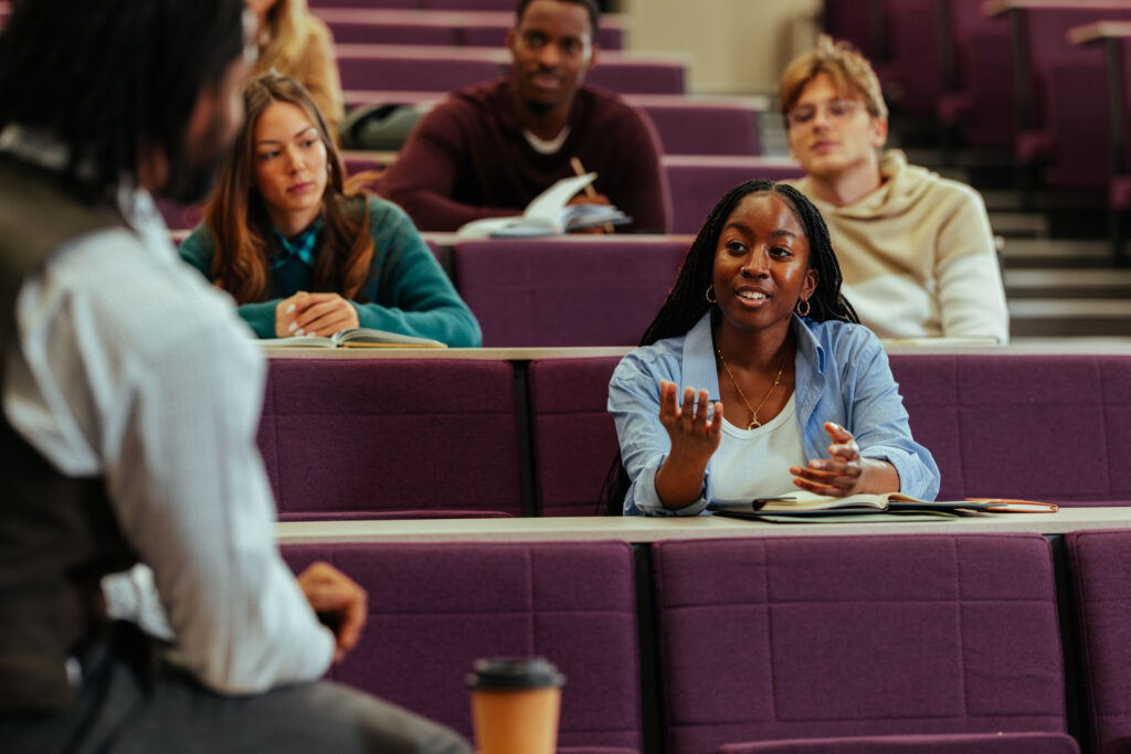 A classroom setting with students sitting in purple chairs. One student in the foreground is gesturing with a hand raised, appearing to ask a question or make a point. Other students are seated behind, some taking notes or reading. A coffee cup is placed on the desk in front of the gesturing student. The instructor is partially visible on the left side of the image, facing the students.