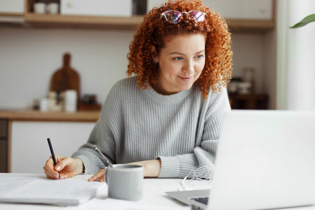 A person with curly hair, wearing a grey sweater, is sitting at a table in a kitchen. The person's face is blurred. They are writing on paper with a pen and have an open laptop in front of them. A grey mug and white earphones are also on the table.