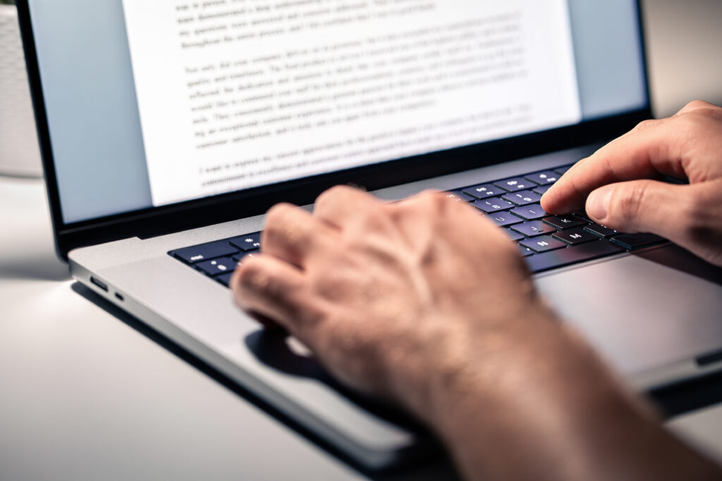 A person is using a laptop with a document open on the screen. The document contains several paragraphs of text. The person's hand is blurred in the foreground, making it difficult to see specific details of what they are doing. The laptop is placed on a white surface.