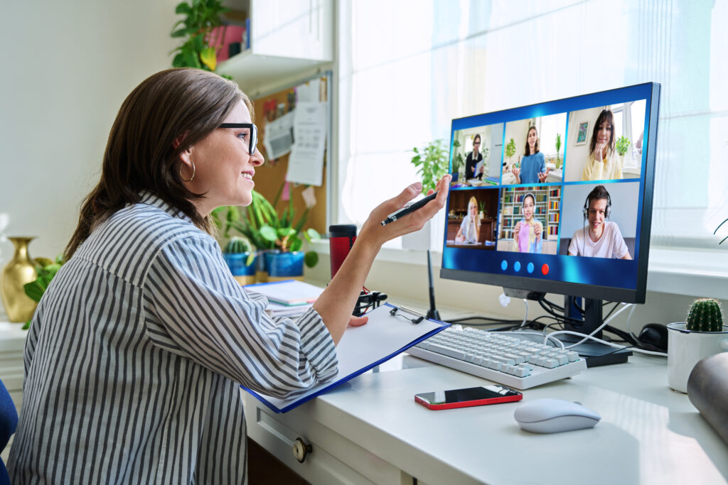 A person is sitting at a desk in front of a computer, participating in a video conference with six other people displayed on the screen. The person is holding a pen and gesturing with their hand while looking at the monitor. The desk has various items including a keyboard, mouse, smartphone, notepad, and some small plants. There are also some papers pinned to a corkboard on the wall behind the person.