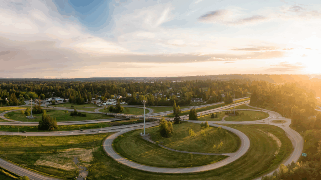 Aerial view of Highway 1 in Fraser Valley, British Columbia during a spring sunset, showcasing a highway interchange surrounded by green fields, trees, and nearby residential areas under a warm, partly cloudy sky.