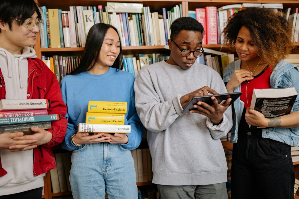 Four people standing in front of a bookshelf filled with books. The person on the left is wearing a red jacket and holding several books, including 'Deutsche Grammatik' and 'Mathematik.' The second person is wearing a blue sweater and holding three yellow books titled 'Deutsch Englisch.' The third person is wearing a gray sweatshirt and using a tablet. The fourth person is wearing a denim jacket over a red shirt and holding an open book while looking at it.
