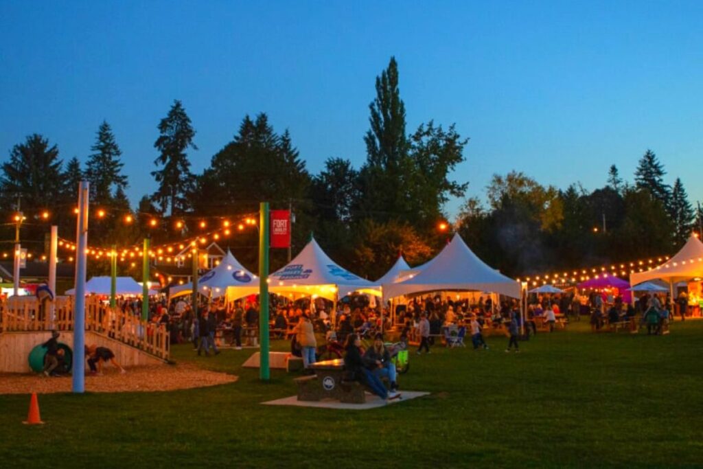Outdoor evening event in Nicomekl, Langley, BC, featuring white tents, string lights, and a lively crowd gathered on a grassy area with children playing in a nearby playground, set against a twilight sky and tall trees.