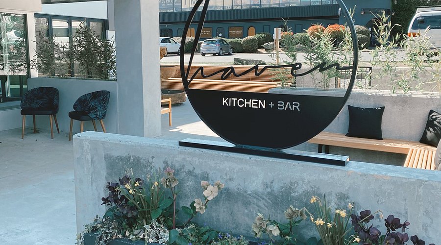 Outdoor seating area at Haven Kitchen + Bar in Nicomekl, Langley, BC, featuring a concrete planter with flowers, cushioned chairs, a wooden bench with pillows, and the restaurant’s name displayed on a circular sign.