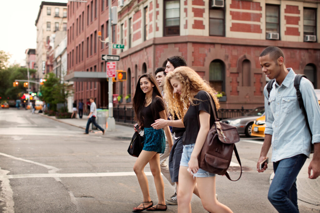 Four young adults cross a city street at an intersection, surrounded by brick buildings and a yellow taxi. They are casually dressed, carrying backpacks or bags, and appear to be engaged in conversation or looking at their phones.