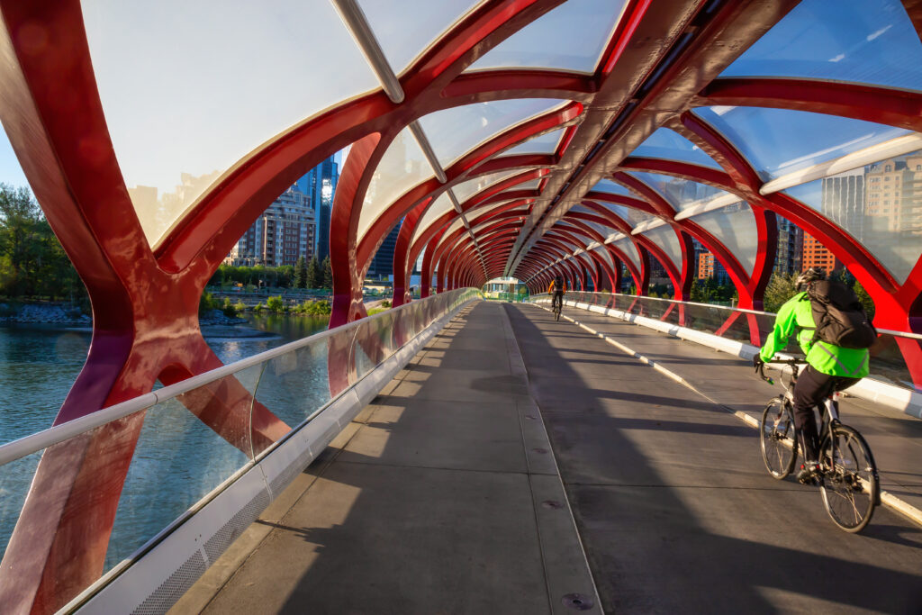 A cyclist in a bright green jacket rides across a modern pedestrian bridge with red arches and glass panels. The bridge spans a river, with city buildings in the background and another person walking ahead. Sunlight casts long shadows on the pathway.