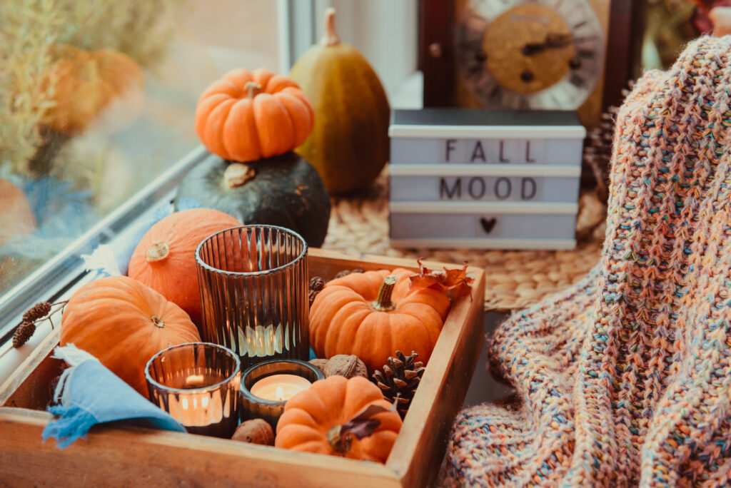 A cozy autumn-themed setup featuring a wooden tray filled with small pumpkins, pinecones, and two lit candles in glass holders. The tray is placed on a woven mat next to a window. In the background, there is a lightbox sign displaying the words "FALL MOOD" with a heart symbol underneath. A knitted blanket with warm colors is draped over the edge of the scene, adding to the cozy atmosphere.