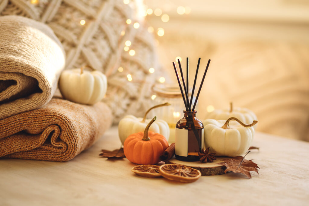 A cozy autumn-themed arrangement featuring a reed diffuser in a brown glass bottle surrounded by small white and one orange pumpkin, dried orange slices, cinnamon sticks, and star anise. The background includes folded knitted blankets in neutral tones and warm fairy lights creating a soft glow.