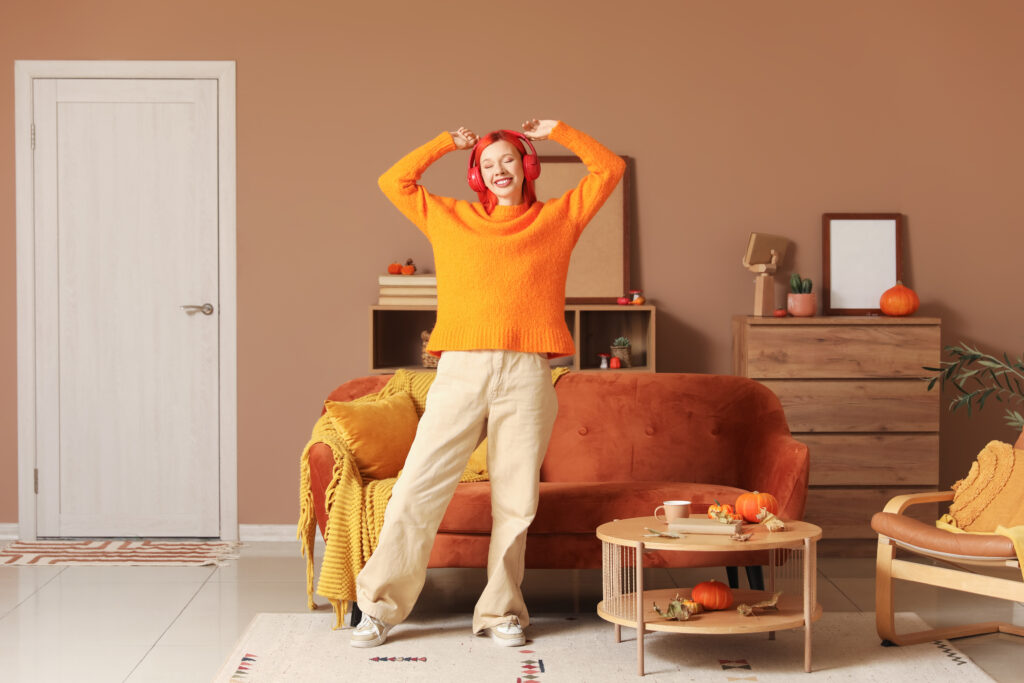 A person in an orange sweater and beige pants stands in a cozy living room with autumn-themed decor, including pumpkins and warm-toned furniture.