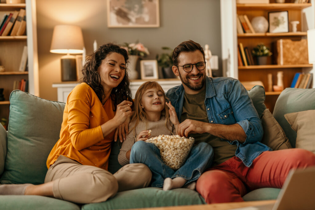 A family of three sitting on a couch in a warmly lit living room, enjoying popcorn together while watching a movie. The parents are seated on either side of their child, who is holding the bowl of popcorn. The room features bookshelves, plants, and soft lighting from a lamp.
