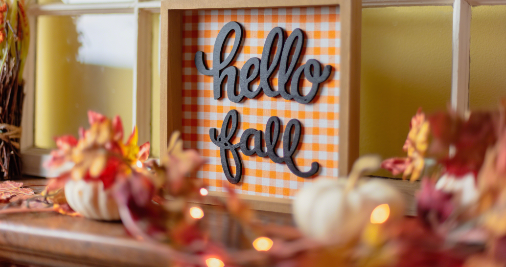 A decorative sign with the words "hello fall" in cursive black letters is set against an orange and white checkered background. The sign is framed in a wooden box and surrounded by autumn-themed decorations, including small white pumpkins and artificial fall leaves. The scene is cozy and festive, capturing the essence of the first day of fall.