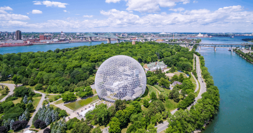 Aerial view of the Montreal Biosphere, a geodesic dome structure surrounded by lush green trees on Saint Helen's Island in Montreal, Canada. The image shows the St. Lawrence River on both sides of the island, with bridges and cityscape visible in the background under a partly cloudy sky.