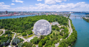 Aerial view of the Montreal Biosphere, a geodesic dome structure surrounded by lush green trees on Saint Helen's Island in Montreal, Canada. The image shows the St. Lawrence River on both sides of the island, with bridges and cityscape visible in the background under a partly cloudy sky.