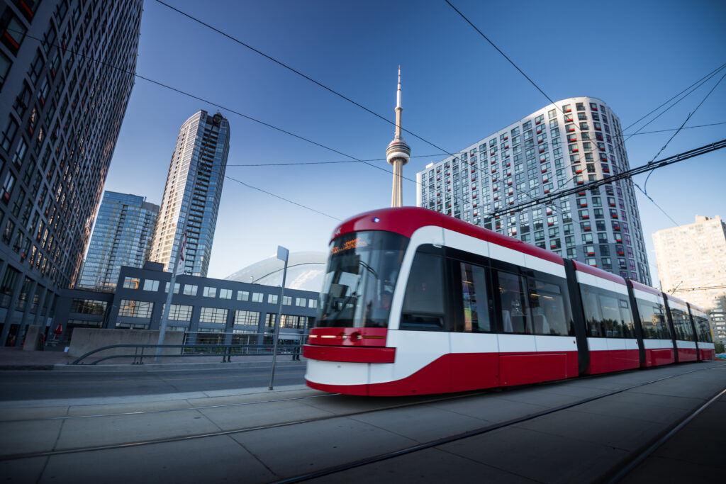 A modern red and white streetcar travels along a track in an urban setting with tall buildings on either side. The CN Tower is prominently visible in the background against a clear blue sky. The streetcar is moving on a concrete path with overhead wires for power.