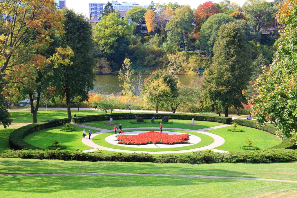 A landscaped garden in High Park, Toronto, featuring a large circular flower bed shaped like a red maple leaf, surrounded by green lawns and walking paths, with people strolling and autumn trees in vibrant shades of green, yellow, and orange in the background near a pond.