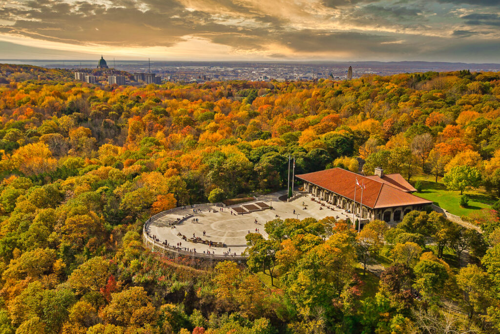 Panoramic view of Mount Royal Park in Montreal during autumn, featuring vibrant fall foliage in shades of orange, yellow, and green surrounding the Kondiaronk Belvedere lookout and pavilion, with people gathered on the terrace and the city skyline visible in the background under a dramatic sunset sky.