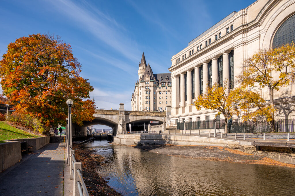 Autumn view of the Rideau Canal in downtown Ottawa, featuring calm water bordered by stone walkways, vibrant fall trees with orange and yellow leaves, and historic architecture including the Château Laurier and a neoclassical building under a clear blue sky.