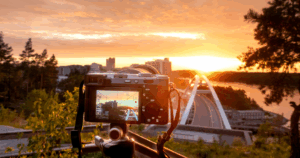 A camera on a tripod captures a scenic sunset over a cityscape, with a bridge and buildings in the background. The sky glows in warm orange tones as the sun sets, and trees frame the view on both sides.