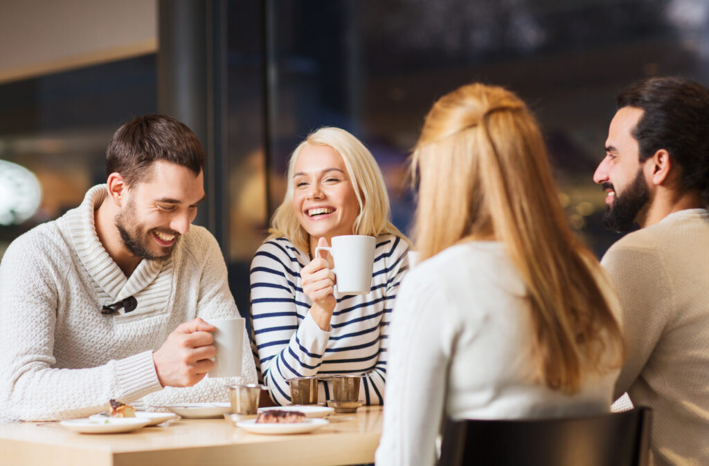 Four people sitting at a table in a café, holding coffee mugs and chatting, with plates and cups on the table.