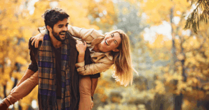 Person carrying another person playfully on their back in a park surrounded by vibrant autumn foliage.