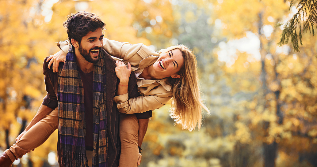 Person carrying another person playfully on their back in a park surrounded by vibrant autumn foliage.